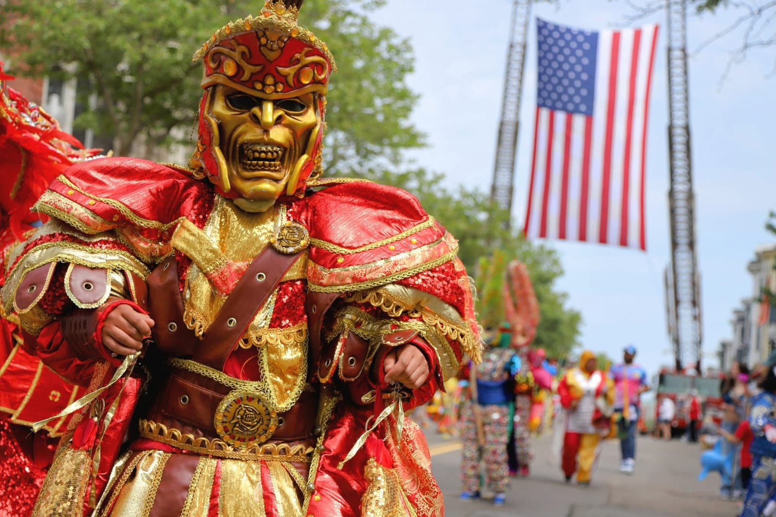 Close-up of a golden warrior carnival mask with red plumes and an American flag hanging behind